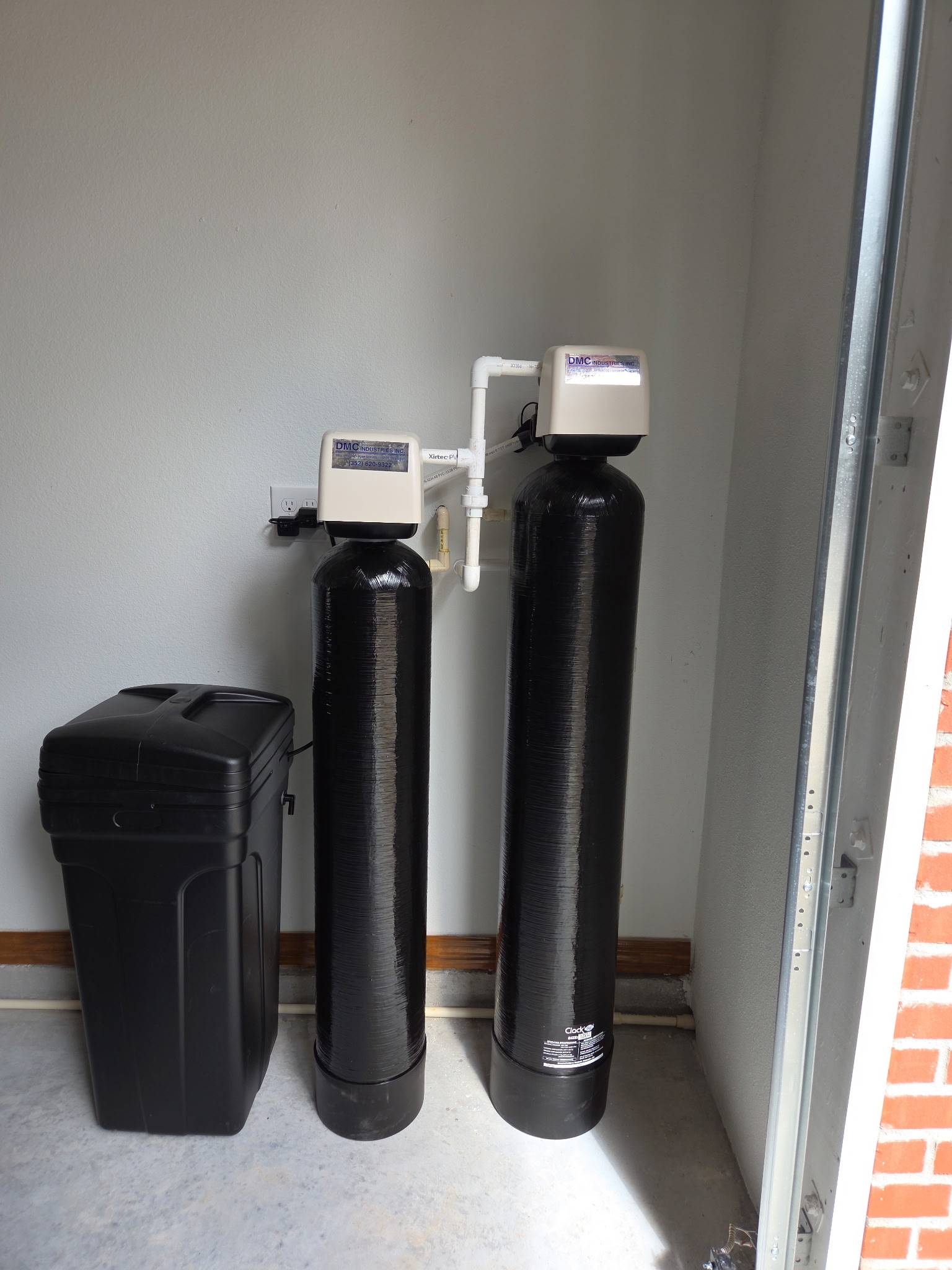 Water filtration system with two black tanks and control equipment mounted on the wall, next to a black trash bin in a garage setting.