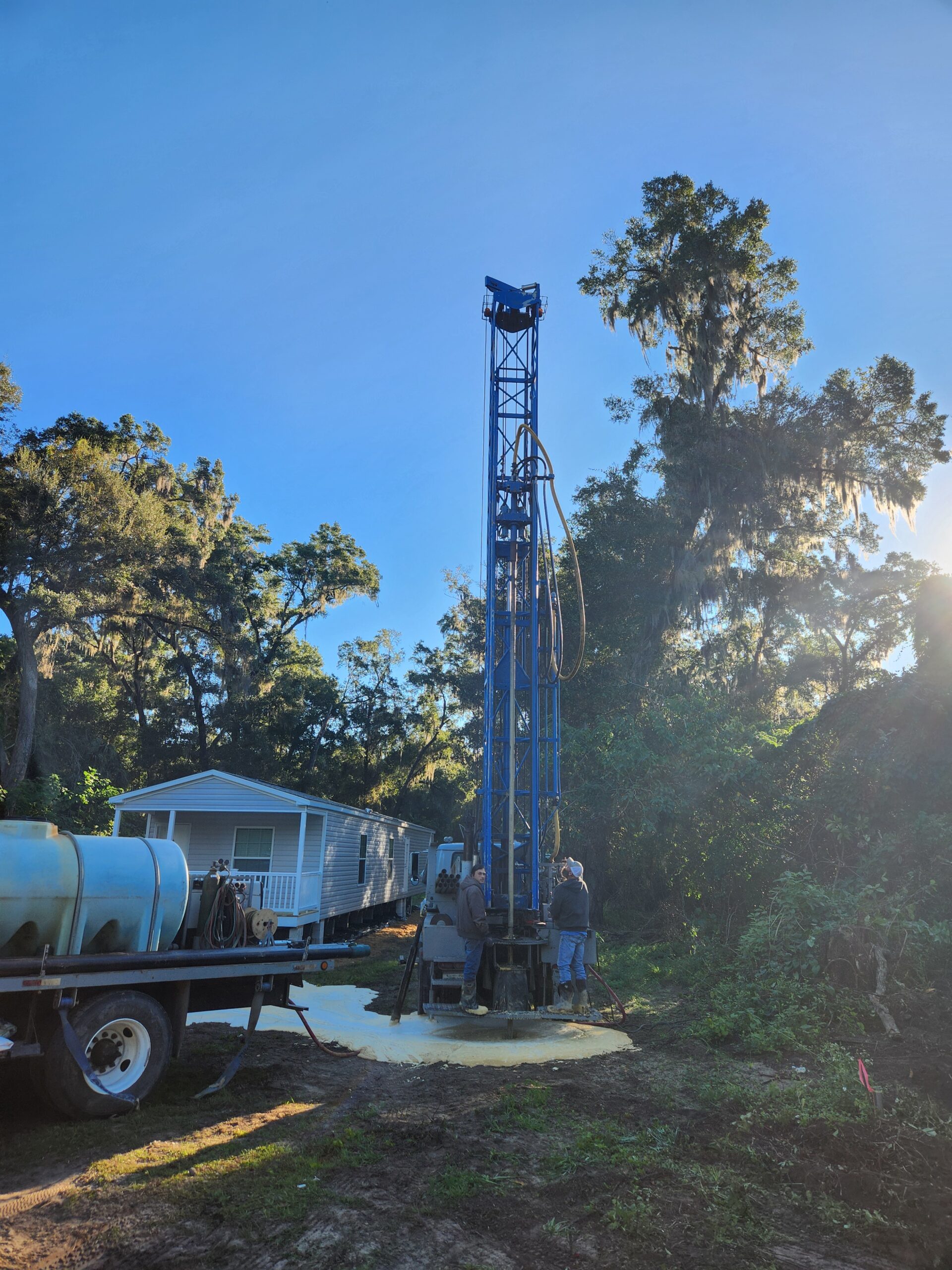 Workers operate a blue drilling rig in a wooded area, with a mobile home nearby. The scene highlights a water well drilling process.